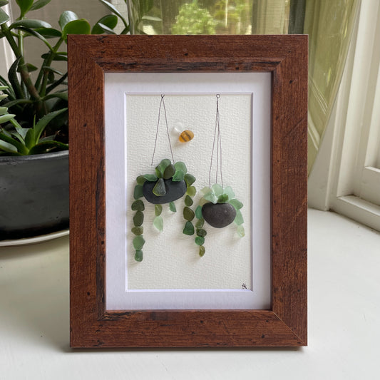 This 5 x 7 inch framed sea glass art picture features two hanging plants made from genuine sea glass, pottery & pebbles with a cute little sea glass bee buzzing about.  Designs may vary. 