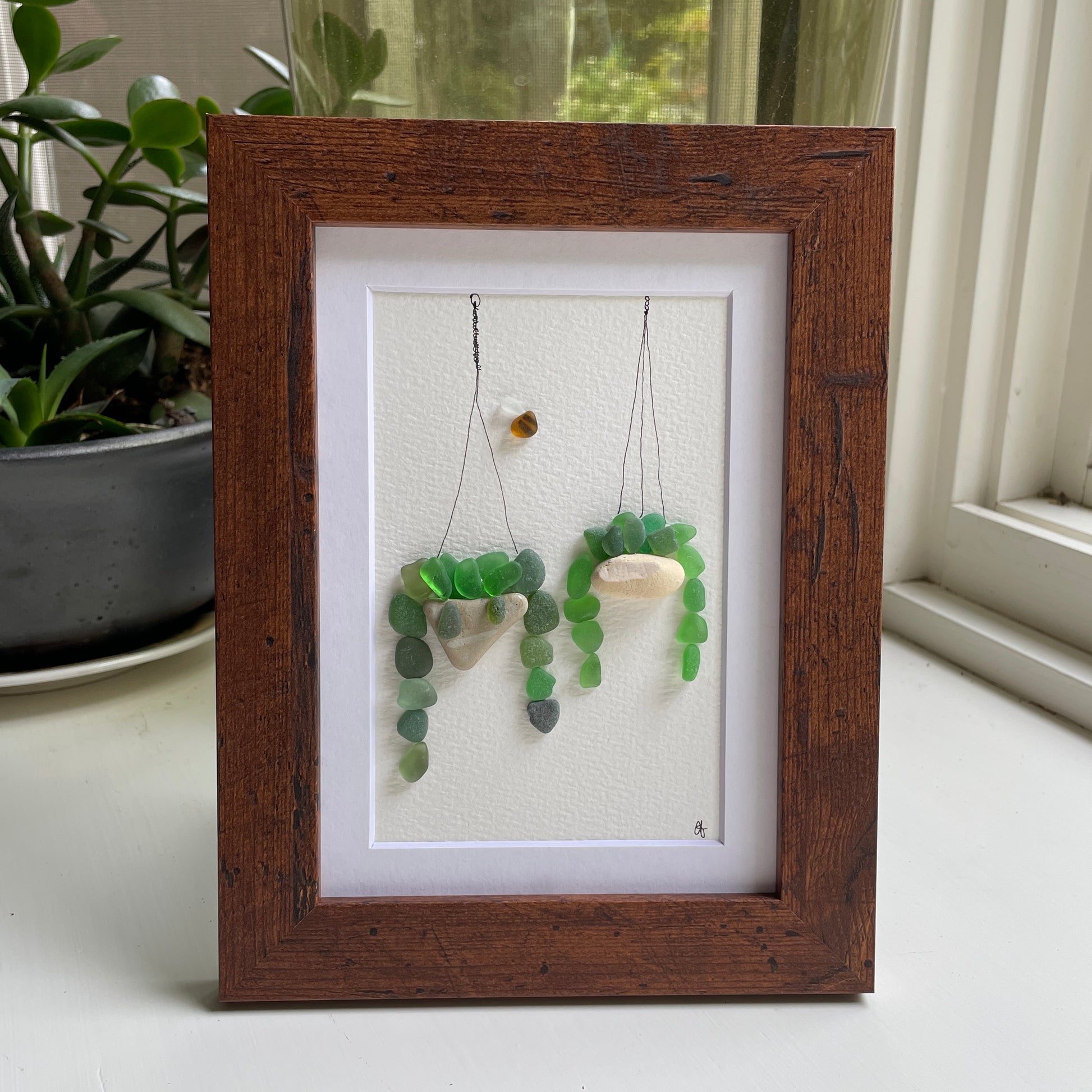 This 5 x 7 inch framed sea glass art picture features two hanging plants made from genuine sea glass, pottery & pebbles with a cute little sea glass bee buzzing about.  Designs may vary. 