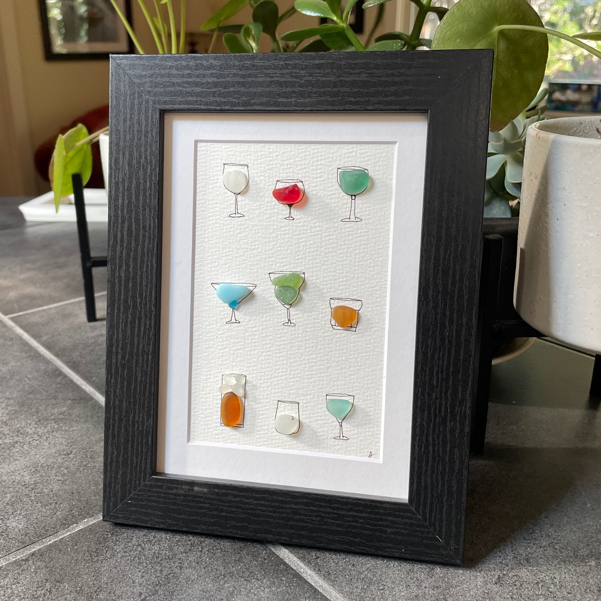 This is a framed 5 x 7 inch sea glass art picture featuring several hand-drawn cocktail and wine glasses that are filled with colorful sea glass. This item is perfect for gift giving and is ready to hang or place on a shelf. 