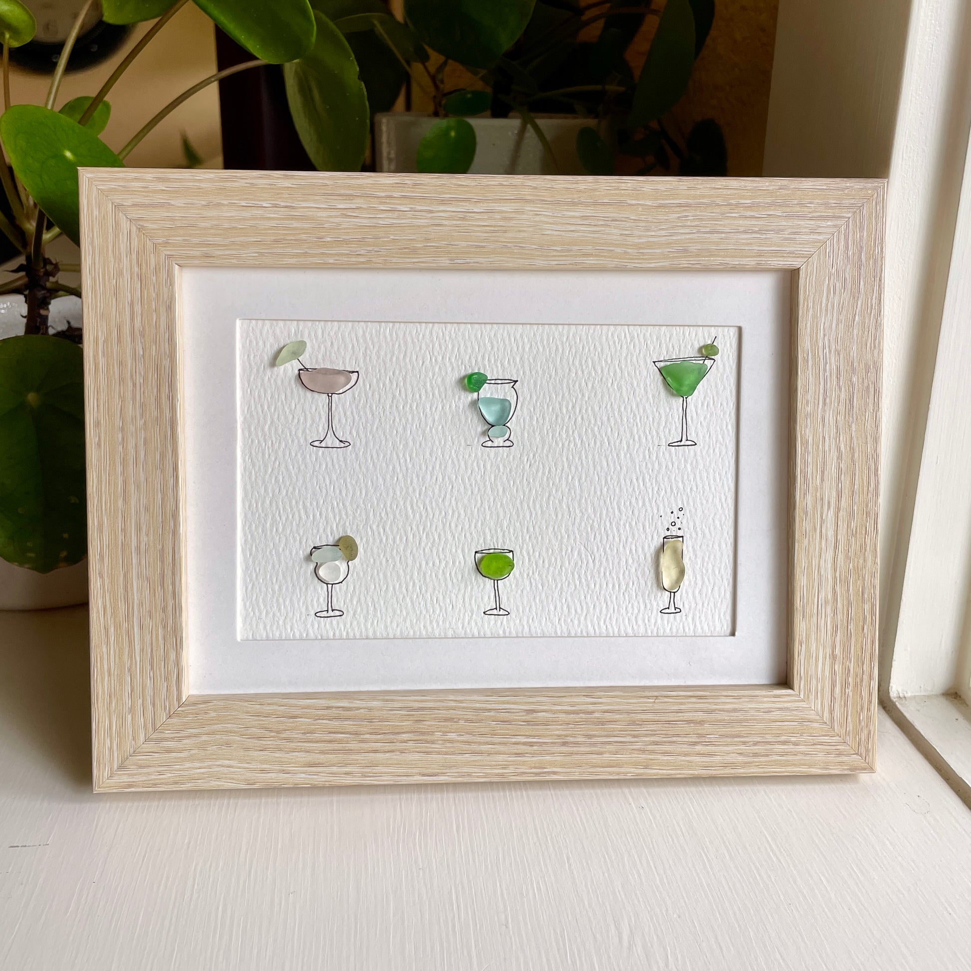 This is a framed 5 x 7 inch sea glass art picture featuring several hand-drawn cocktail and wine glasses that are filled with colorful sea glass. This item is perfect for gift giving and is ready to hang or place on a shelf. 