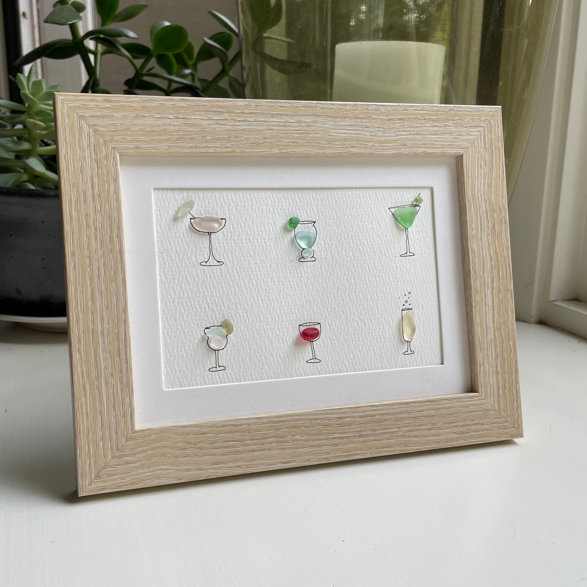 This is a framed 5 x 7 inch sea glass art picture featuring several hand-drawn cocktail and wine glasses that are filled with colorful sea glass. This item is perfect for gift giving and is ready to hang or place on a shelf. 